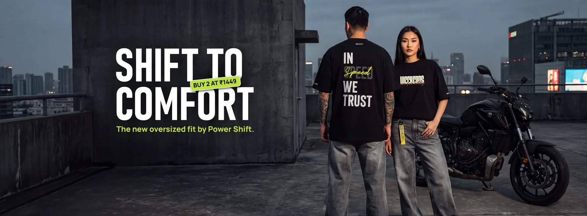 Promotional hero banner for Powershift showcasing a man and woman modeling black oversized graphic t-shirts and gray jeans on an urban rooftop next to a motorcycle. The text reads: "SHIFT TO COMFORT", featuring a promotional badge "BUY 2 AT ₹1449", and the subheadline "The new oversized fit by Powershift."